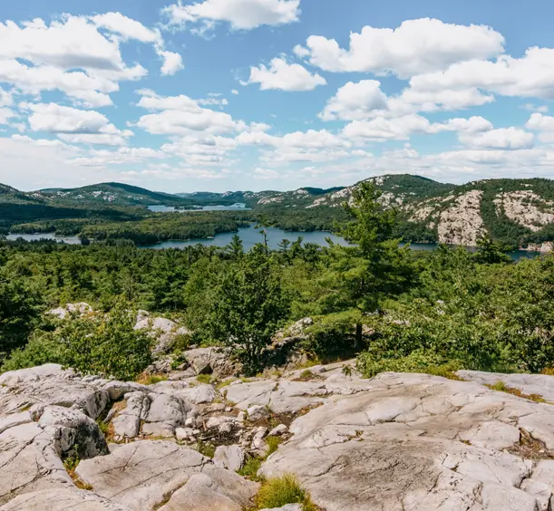 Rocky hilltop view in Killarney Provincial Park overlooking forested valleys, lakes, and white cliffs under a blue sky with scattered clouds.