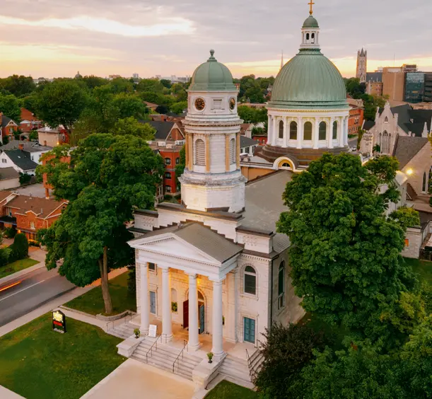 View of St. George’s Cathedral in downtown Kingston, Ontario, with its domed tower rising above surrounding trees under a sunset sky.