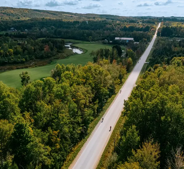 Aerial view of cyclists riding along a rural road in Thornbury, bordered by forest and a winding river on a partly cloudy summer day.