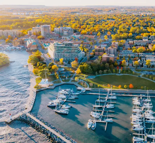 Aerial view of Oakville’s Bill Hill Promenade Park and marina, with sailboats docked and fall trees in the distance amid high and mid-rise buildings.