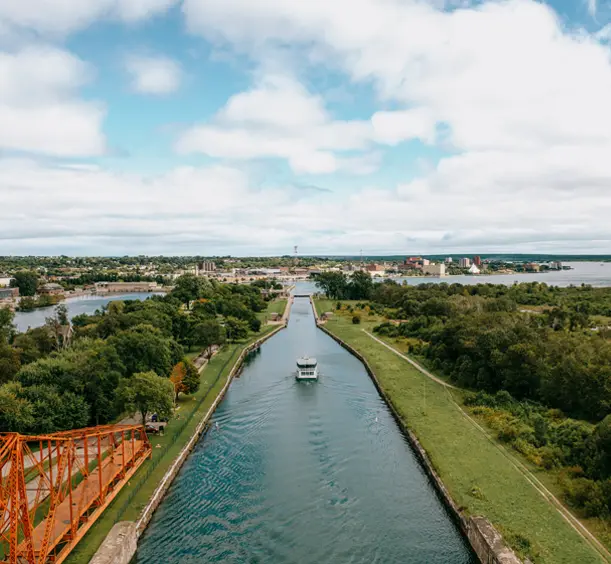 Ein Boot fährt an einem bewölkten Sommertag durch den Sault Ste. Marie Canal und passiert dabei eine rote Drehbrücke mit viel Grün entlang der Küste.