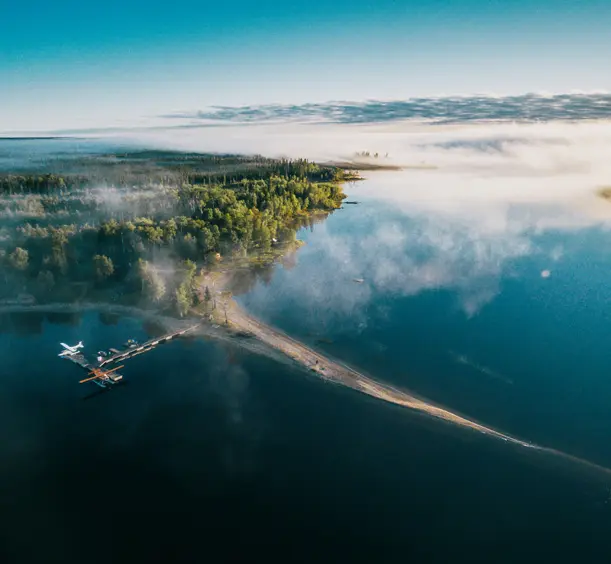Aerial view of Wabakimi Provincial Park, with a docked floatplane, dense green forest, and mist drifting over a still, glassy lake at sunrise.