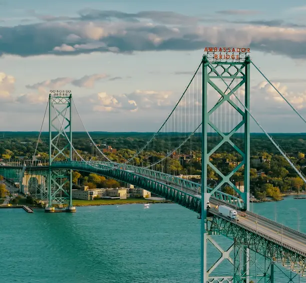 Aerial view of the Ambassador Bridge stretching across turquoise waters, connecting Windsor, Ontario, with the U.S., under a sunset sky.
