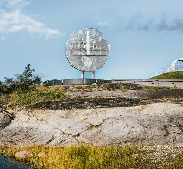 A giant silver-coloured replica of a nickel coin, the Big Nickel, standing above rocky terrain and a pond at Dynamic Earth, Sudbury, on a summer day.