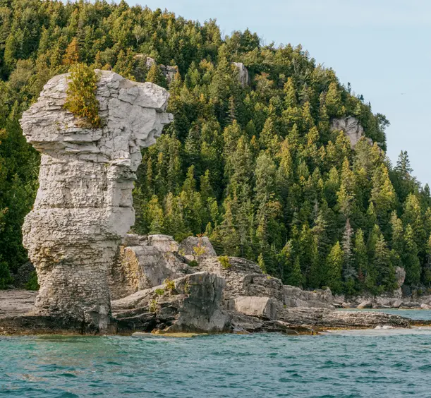 A massive flowerpot rock formation rising from the shoreline of Flowerpot Island in Tobermory, with a forested cliff in the background.