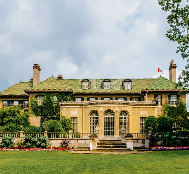 Arrière du domaine Parkwood, avec une grande façade en pierre, des fenêtres cintrées, un toit vert et des jardins soignés, sous un ciel nuageux d’été.