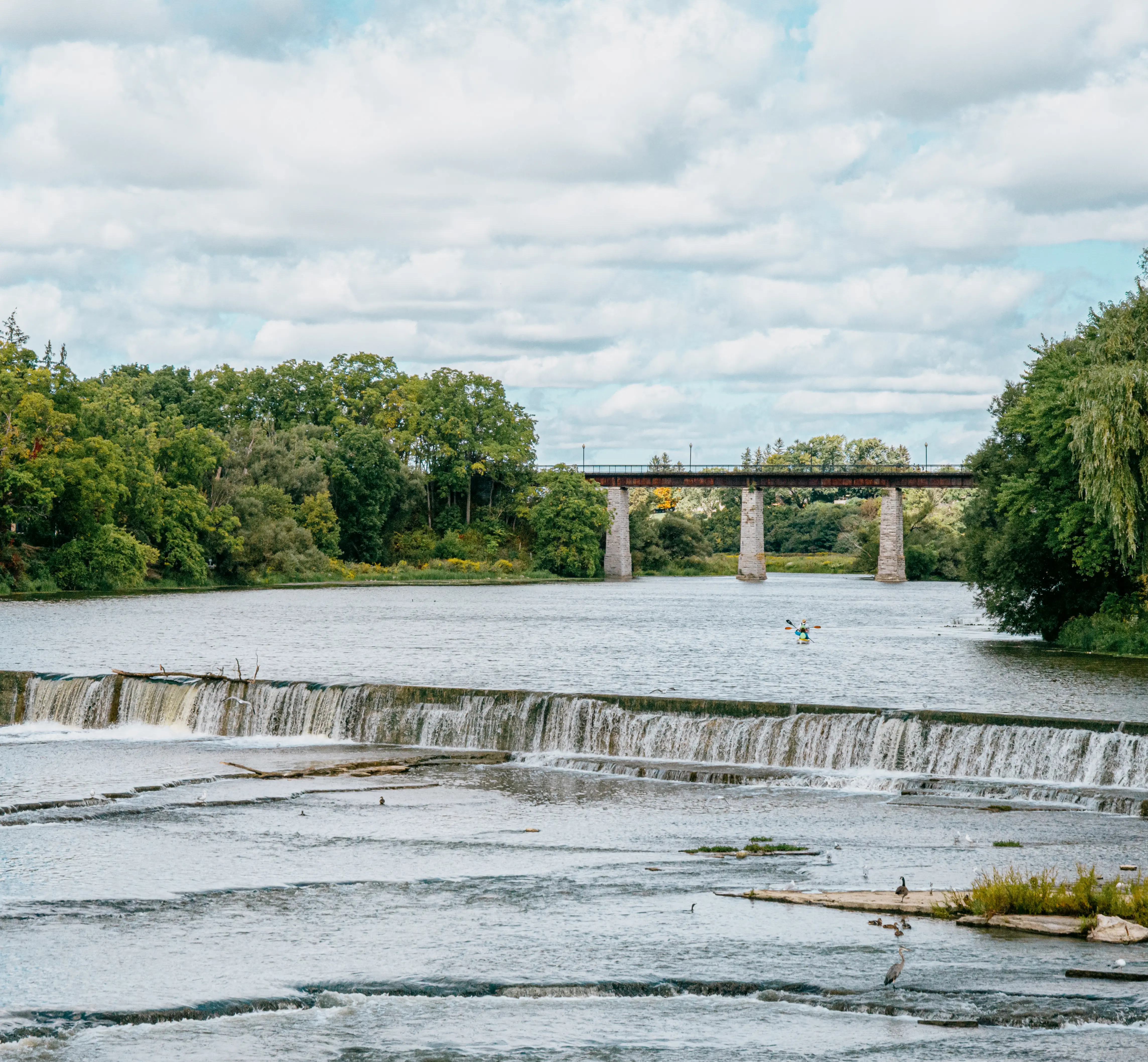 An einem teilweise bewölkten Sommertag stürzt Wasser über einen niedrigen Damm in St. Marys, Ontario, eingerahmt von dichten Bäumen und einer Steinbrücke in der Ferne.