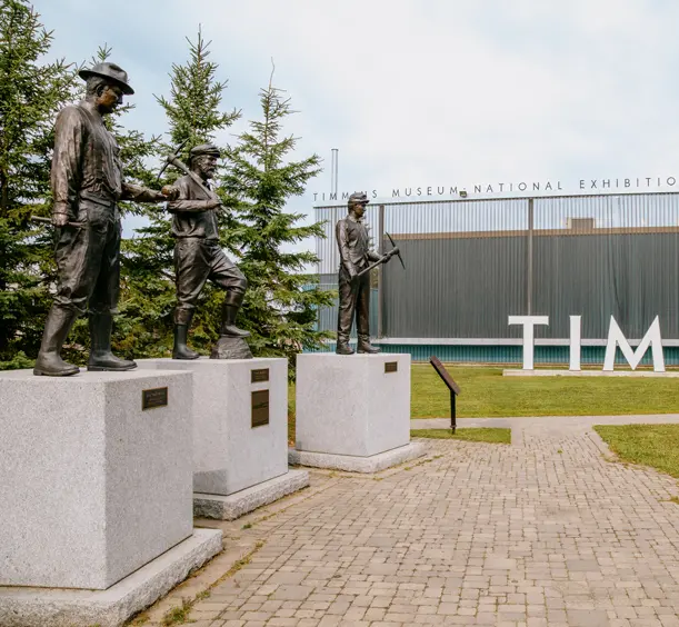 Mining statues outside the Timmins Museum National Exhibition Centre, with large TIMMINS letters in the background on a cloudy day.
