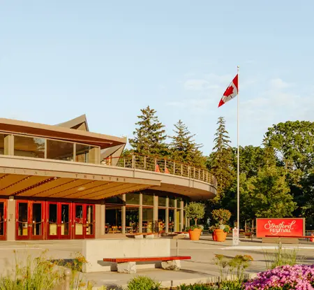 Stratford Théâtre Stratford Festival en été, avec des portes rouges, des drapeaux canadiens et des jardins en fleurs en avant-plan, sous un ciel clair.