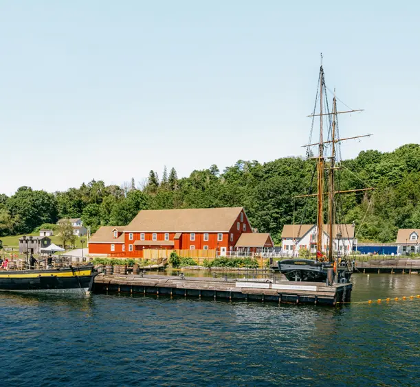 Tall ship docked at Discovery Harbour in Penetanguishene, Ontario, with red heritage buildings and lush greenery under a clear summer sky.