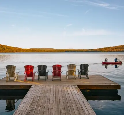 Une rangée de six chaises Muskoka sur un quai fait face à un lac calme avec un canot et les couleurs d'automne au loin.