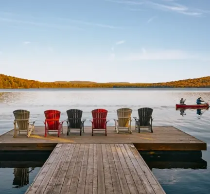 A row of six Muskoka chairs on a dock face a calm lake with canoe and fall colour in the distance.