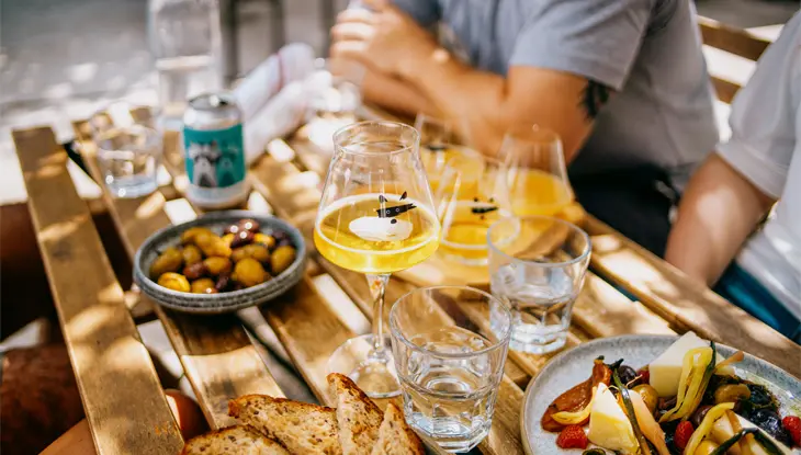 Outdoor dining scene in Toronto with people gathered around a wooden table, enjoying drinks and shared plates of bread, cheese and olives.