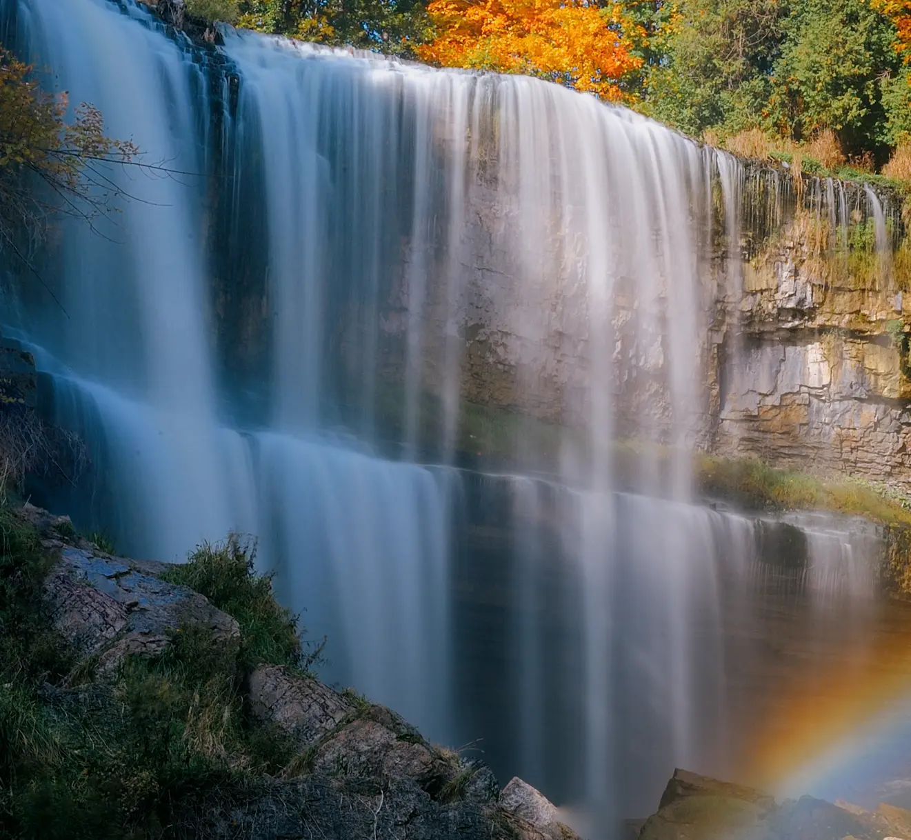 A cascading waterfall over the rocky escarpment surrounded by fall foliage and a rainbow in the foreground.