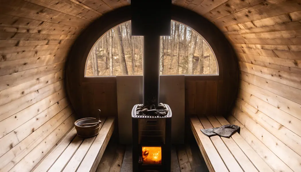 Un cubo y un par de guantes descansan en los bancos junto al fuego de leña y piedras calientes en una sauna de barril de cedro. 
