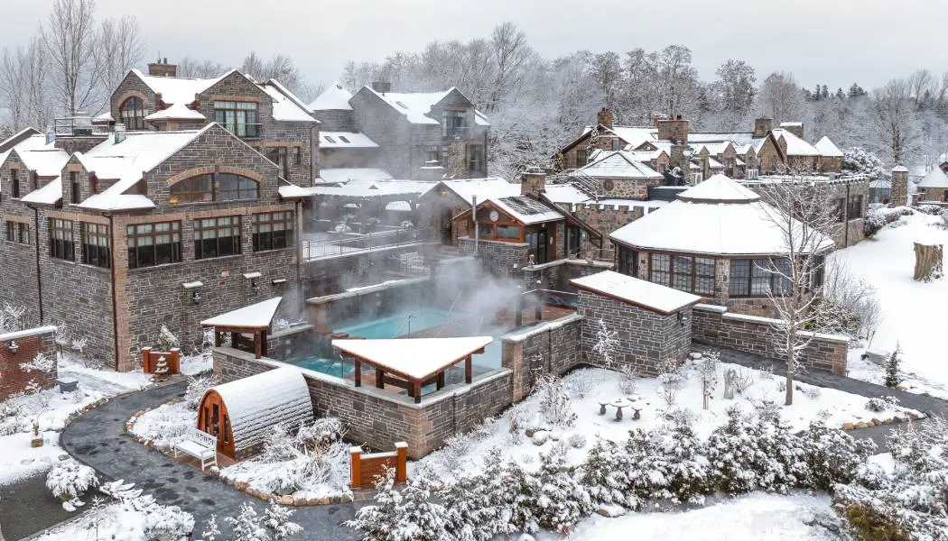 Historische Steingebäude, die mit leichtem Schnee bedeckt sind, umgeben einen beheizten Außenpool, aus dem Dampf aufsteigt – das ist das Ergebnis eines Kurortes.