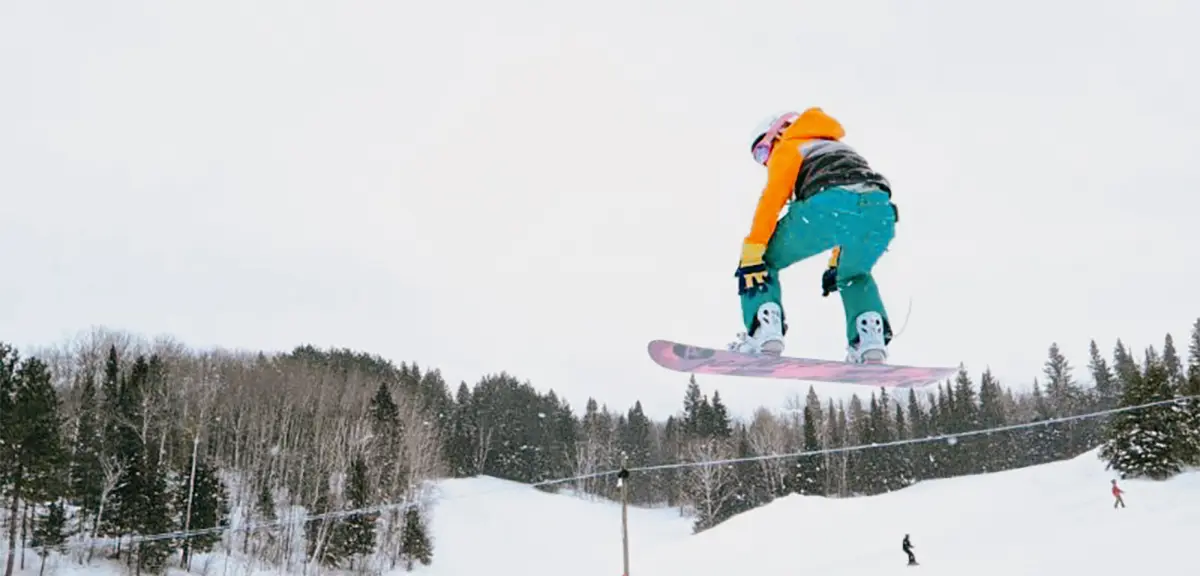 Planchiste dans les airs effectuant une figure sur une pente enneigée, sous un ciel nuageux, avec une forêt au loin.