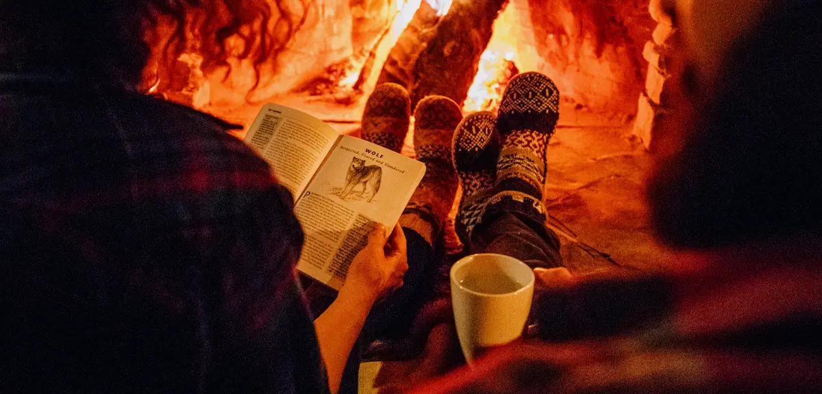 Two people sit side by side in front of a lit fireplace, wearing cozy socks; one reads a book while the other holds a mug, creating a warm, intimate winter atmosphere.