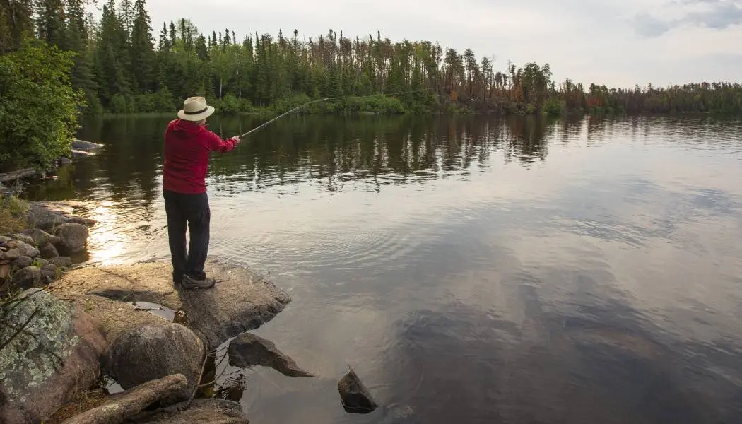 A man standing on a rocky shore casts out a fishing line into a calm lake in an Ontario provincial park.