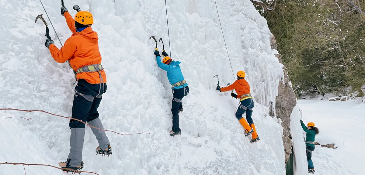 ヘルメットと冬装備を身に着けた4人が、雪に覆われた森の中で、ロープとピッケルを使って凍った壁を登ります。
