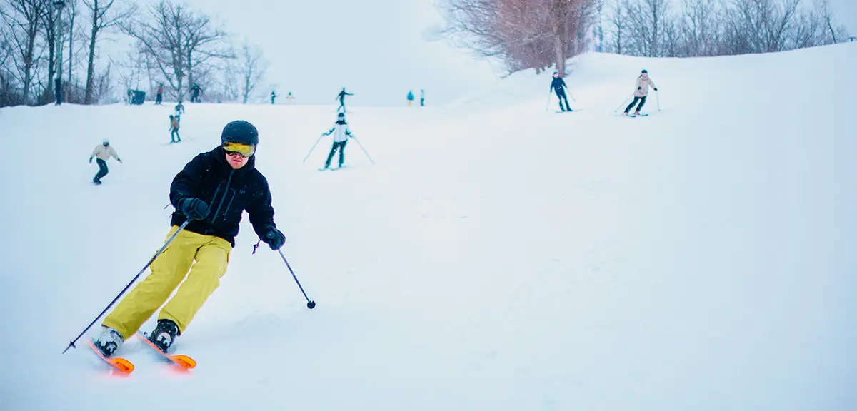 霧の日に、黄色いパンツをはいたスキーヤーが雪に覆われた斜面を滑降し、背景には他のスキーヤーが見える。