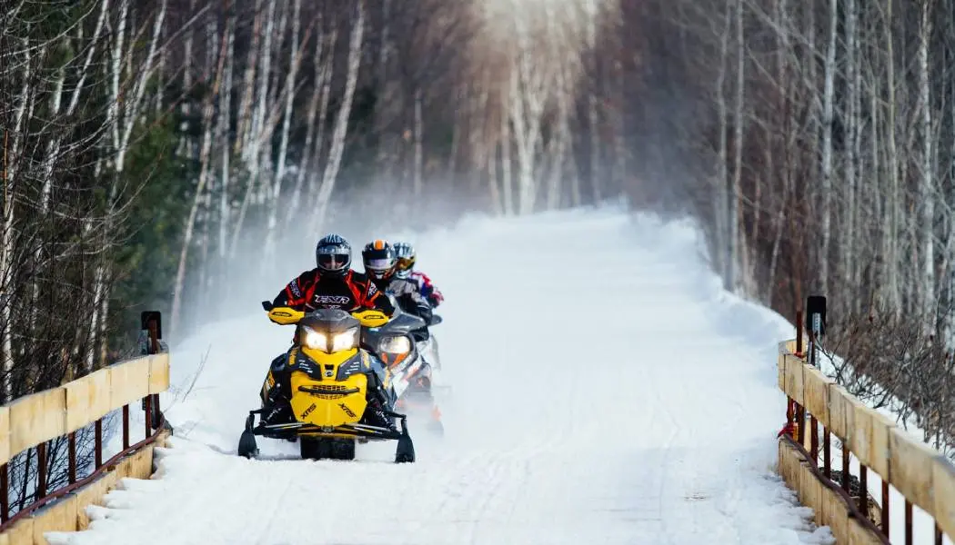 Una fila de tres conductores de motos de nieve conduciendo sus trineos a través de un puente cubierto de nieve rodeado de bosque al fondo.