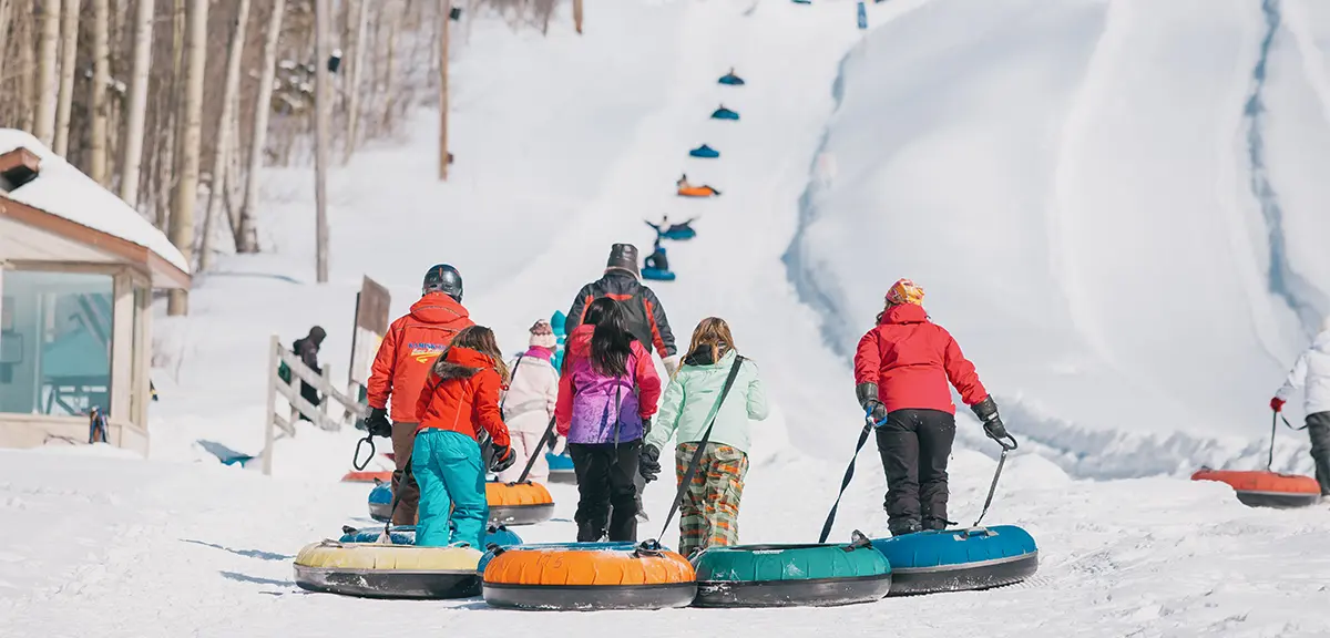 Las familias tiran coloridos tubos de nieve cuesta arriba en un parque de tubing, con pistas nevadas y árboles de fondo.