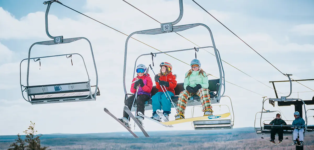 Three people ride a ski lift with snowboards and skis, heading up a snowy slope on a clear winter day.