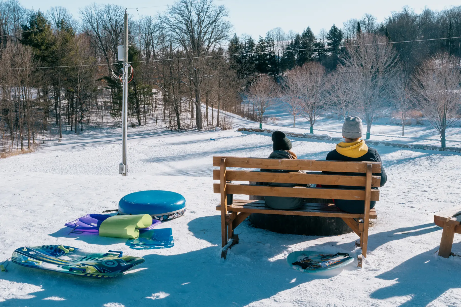Deux personnes assises sur un banc au pied d’une colline enneigée bordée d’arbres, avec des traîneaux et des chambres à air à côté du banc.