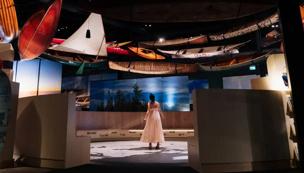 한 여성이 캐나다 카누 박물관의 비공개 투어에 참여하여 전통 카누와 카약을 포함한 다양한 수상 운송 수단들을 둘러보고 있다.