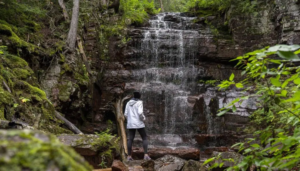 A hiker stops to admire a gentle waterfall surrounded by mossy rocks, trees and lush greenery in Northern Ontario.