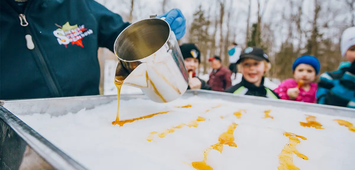 Fresh maple syrup poured onto snow during an outdoor winter festival, with children watching in the background.