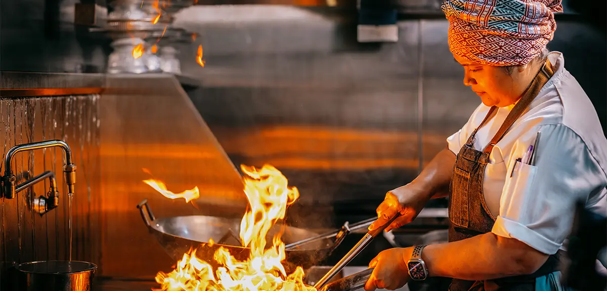 A woman chef expertly flame-cooks a dish in a large wok in the kitchen of Kiin, a MICHELIN-starred restaurant in Toronto.