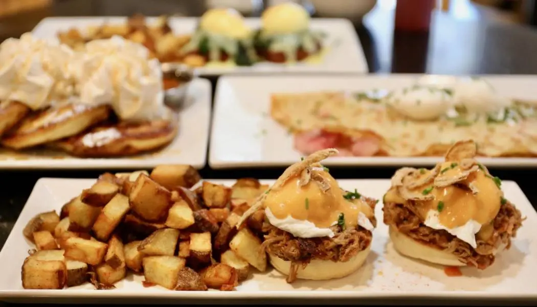 Un assortiment de plats de brunch gastronomiques servis sur des assiettes blanches, comprenant des œufs Bénédicte au porc effiloché, des pommes de terre croustillantes et des crêpes à la crème fouettée.