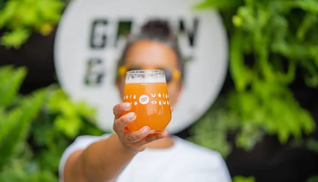 Eine Frau hält ein frisch gezapftes Glas Craft-Bier hoch und steht vor dem Brauereischild.