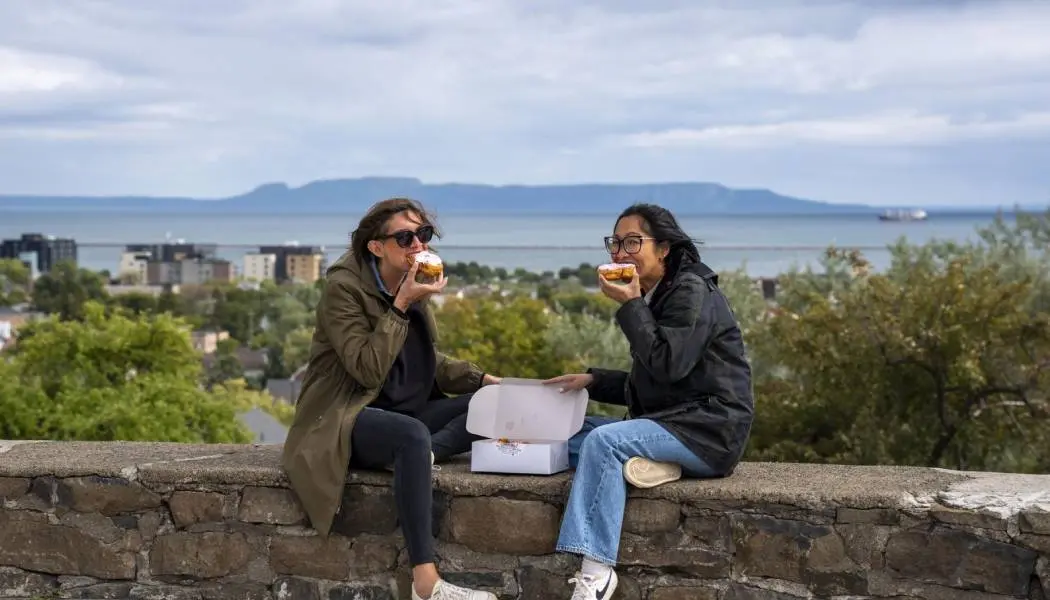Dos mujeres disfrutan comiendo pasteles persas en Thunder Bay, con el Sleeping Giant de fondo.