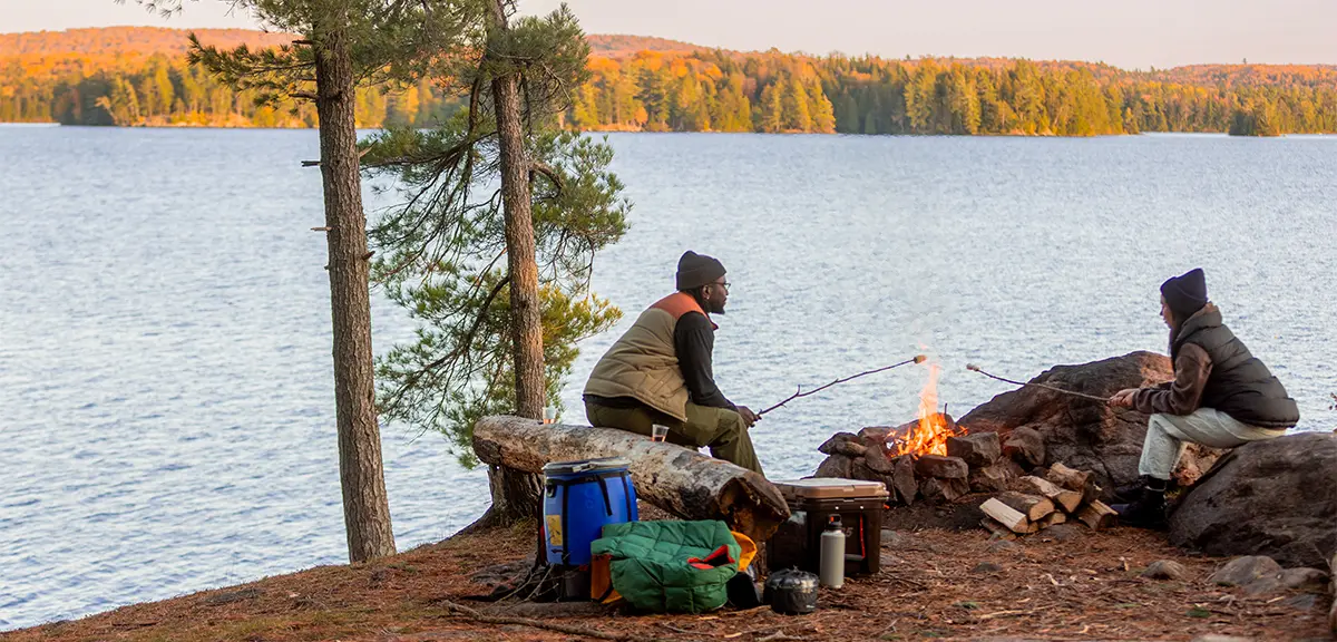 Dos personas sentadas junto a una fogata en la orilla rocosa de un lago, asando malvaviscos con el equipo de acampada cerca.