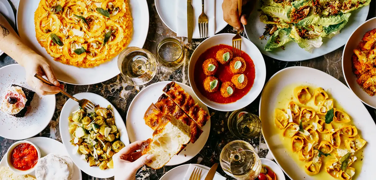 A top view of a table filled with vegan shared plates including pasta, risotto, salad and bread with hands reaching in to eat.