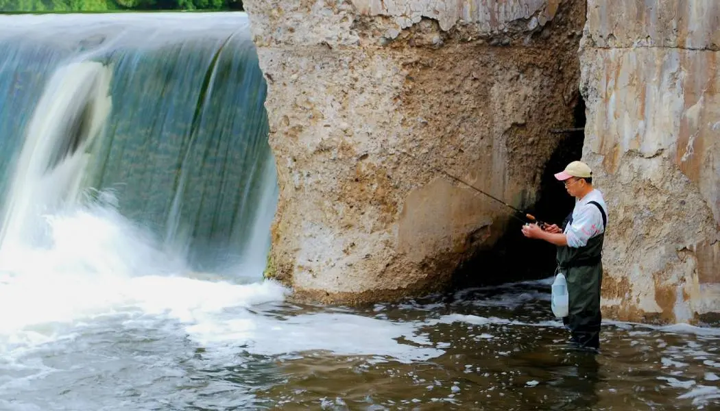 A man wearing waders is fly fishing beside a waterfall at the Paris Dam in Brant County.