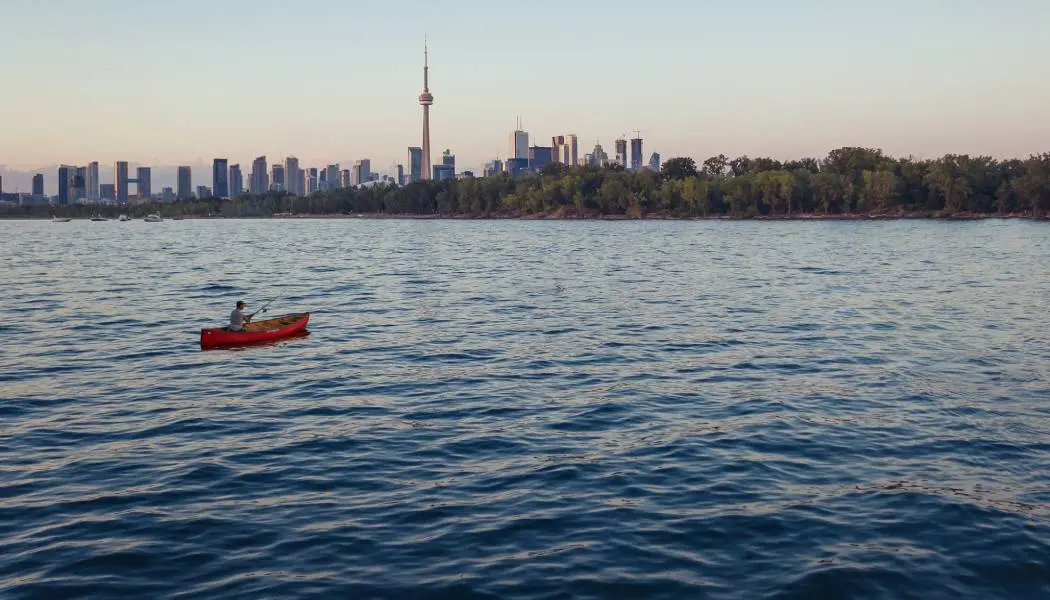 Un homme lance sa ligne de pêche depuis un canoë dans les eaux du port de Toronto, avec la Tour CN et la silhouette de la ville en arrière-plan.
