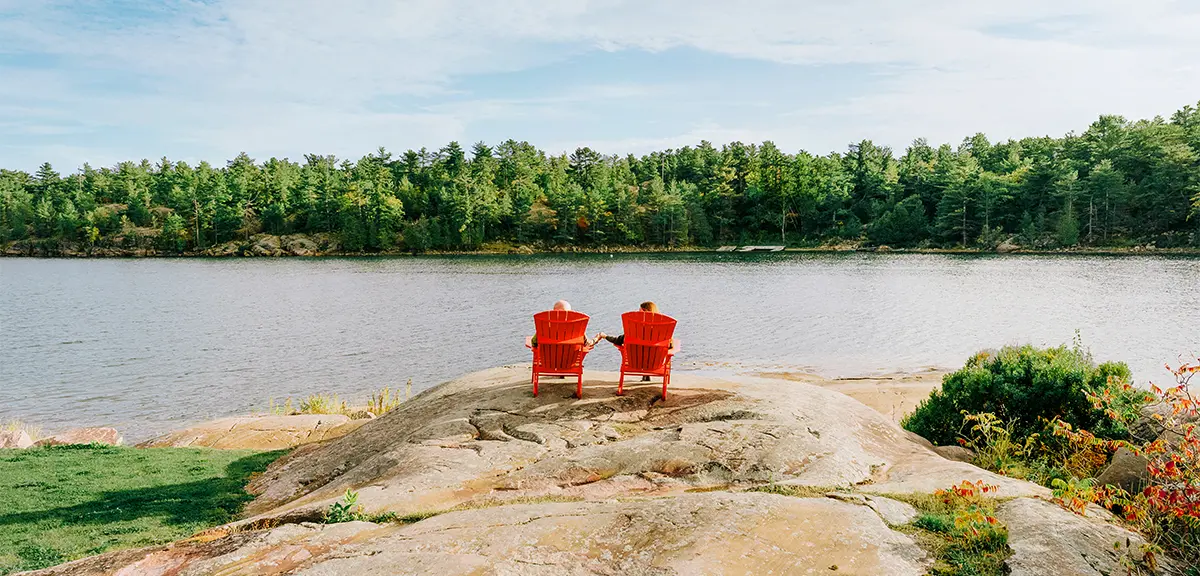 Two red chairs on a rocky shoreline overlooking a calm lake surrounded by forest.