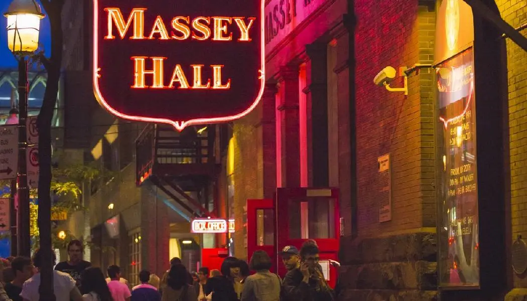 Die leuchtende Neonreklame für die Massey Hall hängt über einem Bürgersteig, der abends von Fußgängern und Konzertbesuchern bevölkert ist.