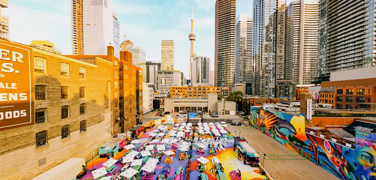Foule rassemblée sur une terrasse extérieure dotée de tables de pique-nique et de parasols, avec la ville en arrière-plan.