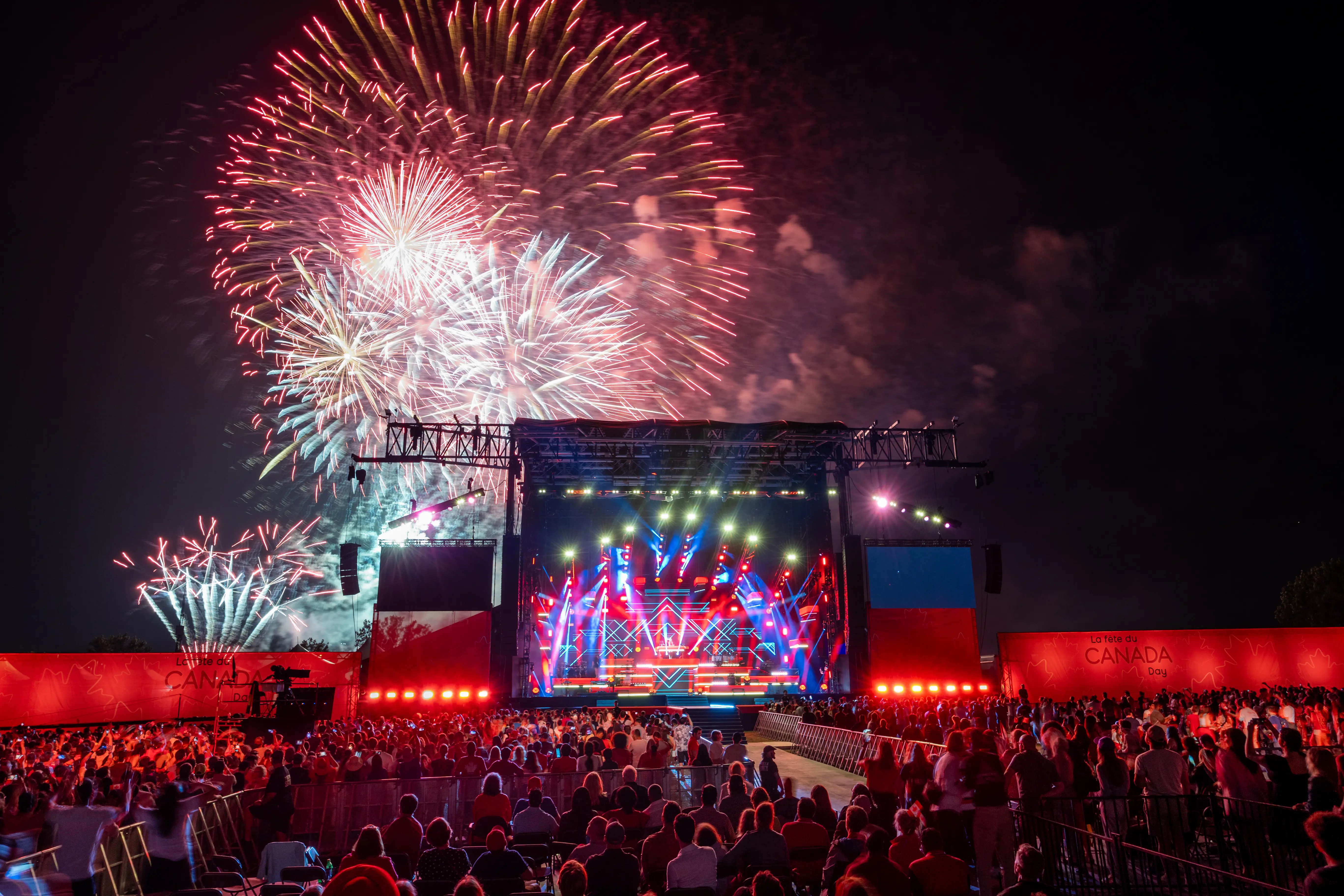 Thousands of attendees standing in front of the Canada Day show stage at Parliament Hill in Ottawa, while illuminated with strobes of light and fireworks