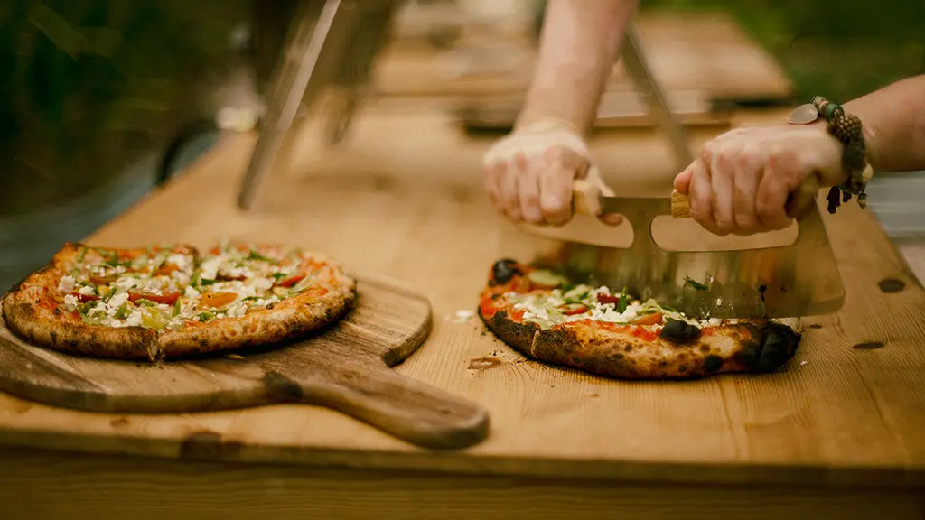 Zwei frisch gebackene, im Holzofen gebackene Pizzen liegen auf einer Holzplatte. Eine Pizza wird gerade mit einem Mezzaluna geschnitten.
