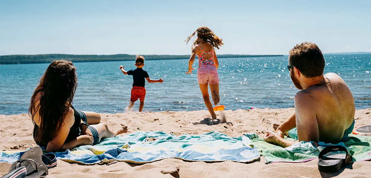 Two adults relax on towels at a sandy lakeside beach while two children run toward the water on a sunny day with clear blue sky.  