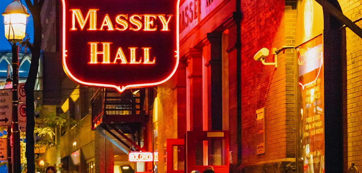 Orange and pink lit Massey Hall sign hangs above a group of people at dusk.