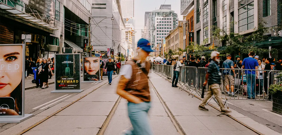 Pedestrians cross a downtown streetcar corridor lined with tall buildings, crowds and advertising boards.