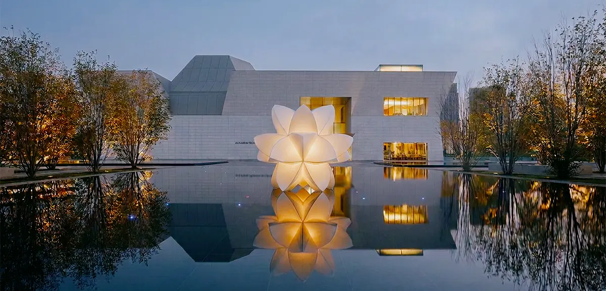 Musée blanc avec une fleur lumineuse reflétée dans un bassin, entouré d’arbres aux couleurs d’automne.