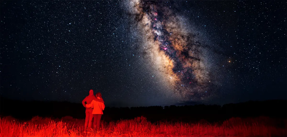 Two people stand together in a grassy field illuminated by red light, gazing up at the star-filled night sky and the bright Milky Way.